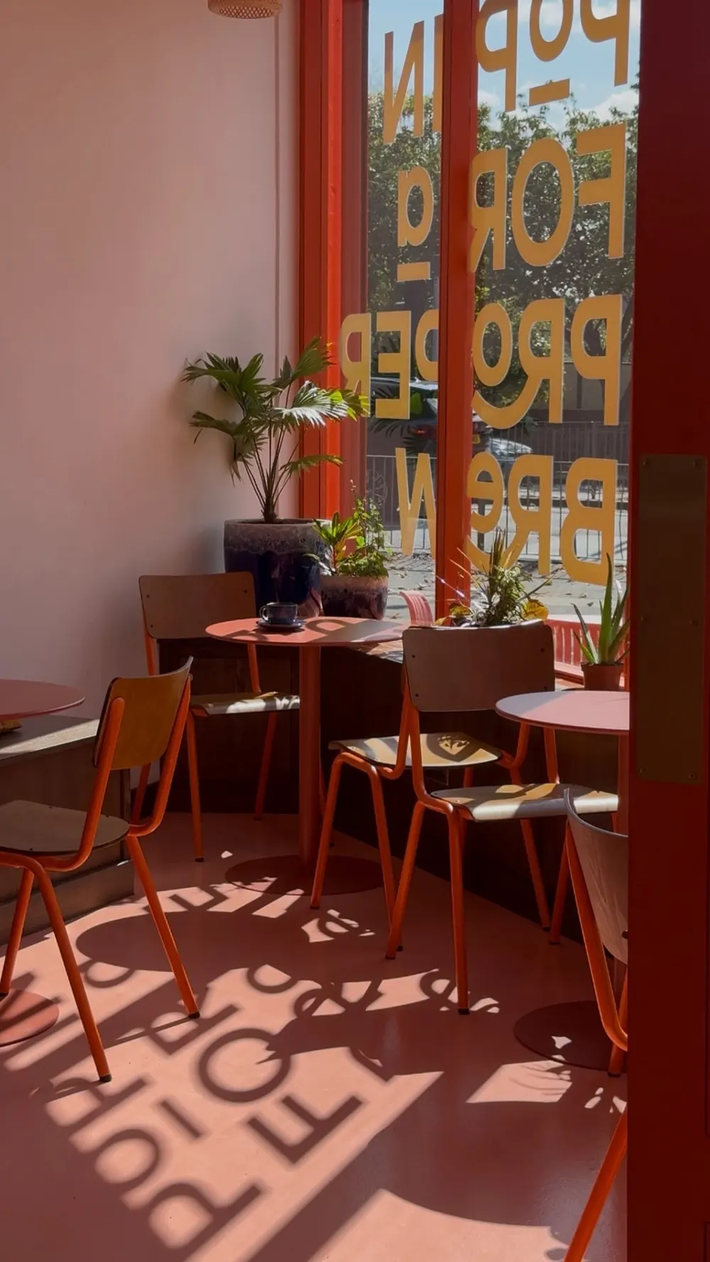 Sunlight through the Pop in for a proper brew window lettering, across orange chairs and tables